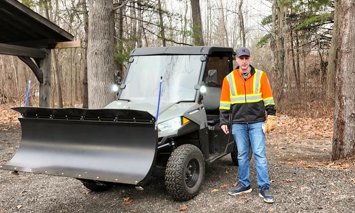A man in a safety vest stands beside a black utility vehicle with a snow plow attachment, highlighting its versatility.