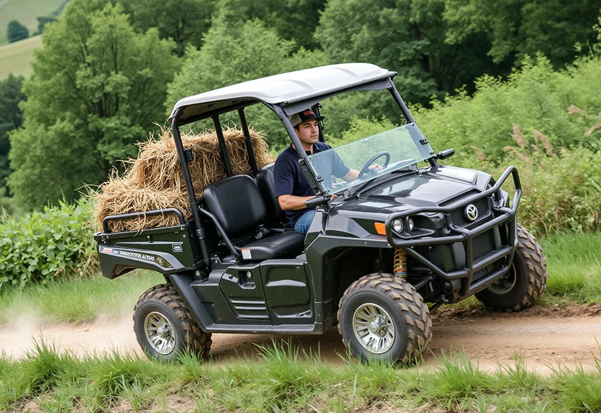 A man driving a black utility vehicle loaded with hay bales on a dirt path, showcasing golf cart battery capabilities.