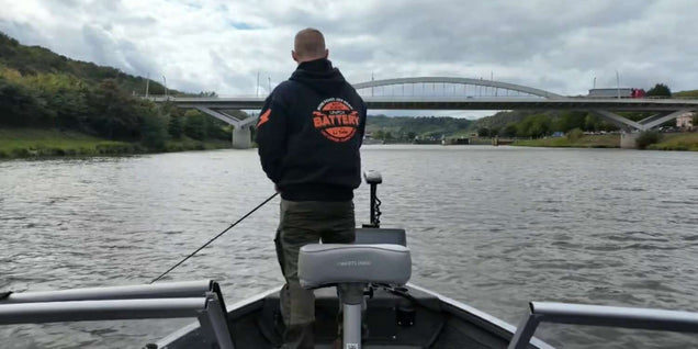 Man fishing from the bow of a boat on a river, with a bridge in the background and a trolling motor